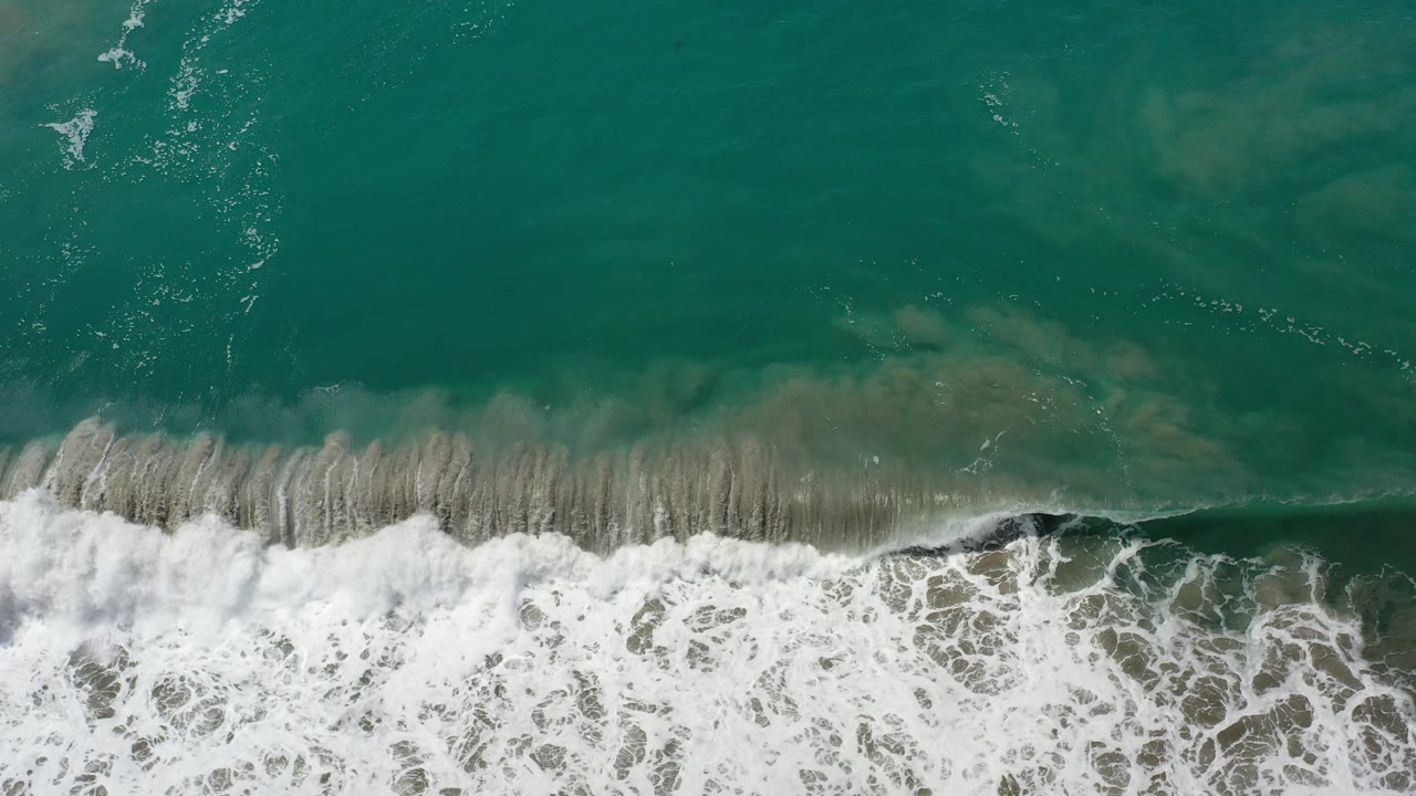 Overhead view of teal water and a white surf band reaching the sandy shoreline in Los Cabos, Mexico, with no visible people.