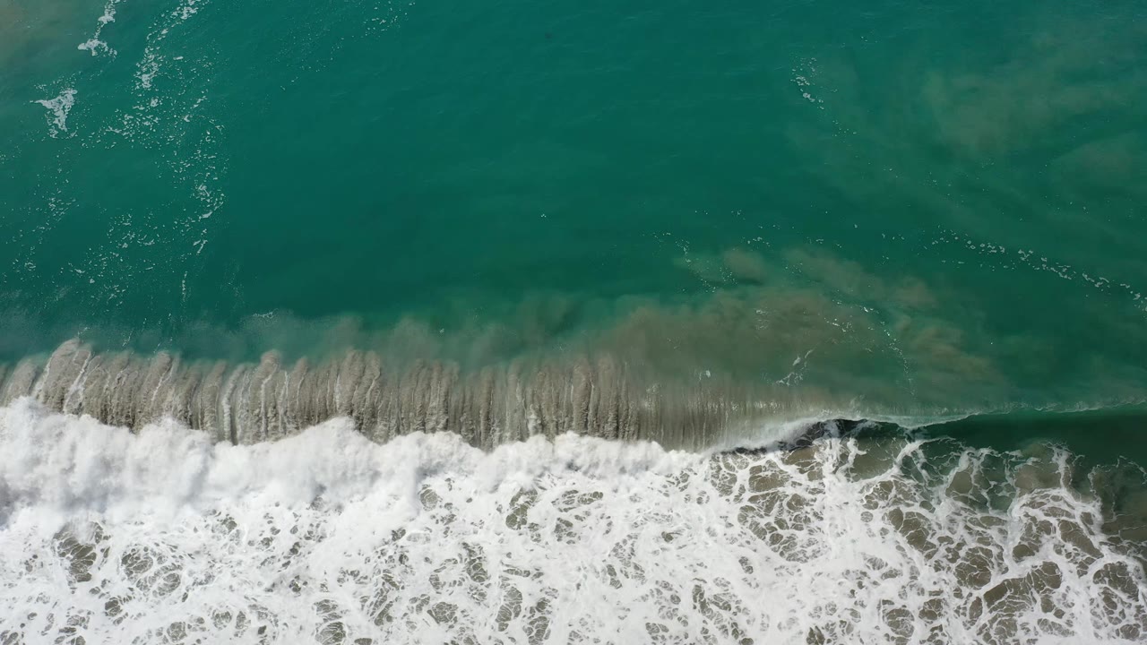 A fresh line of white foam spreads along the lower frame while teal Los Cabos water and submerged sand fill the upper area, with no visible people.