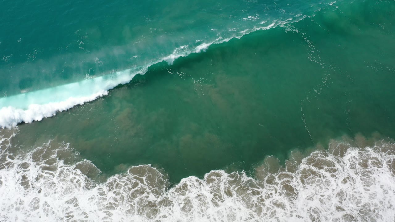 Overhead view of green ocean water in Los Cabos, Mexico with a white breaker entering from the upper left and foamy wash over rocks below, no visible people.
