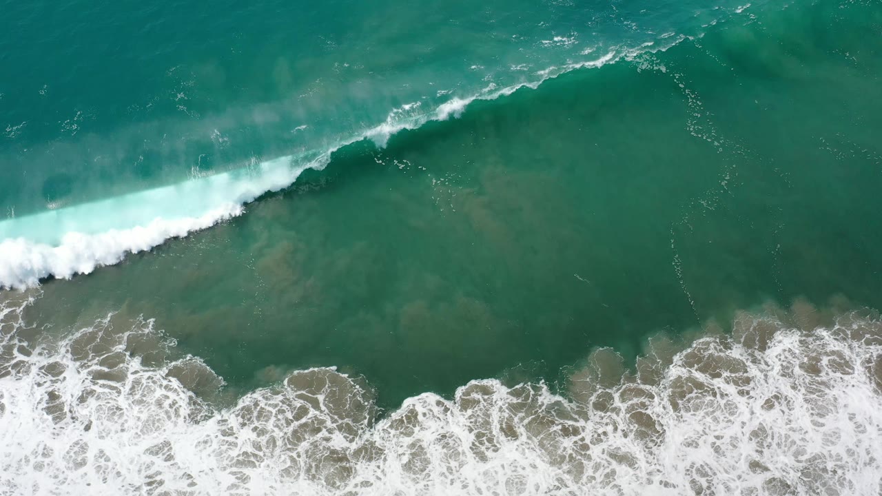 Overhead Los Cabos surf frame with the white wave crest farther across the green water and lace-like foam gathering along the rocky lower edge, no visible people.