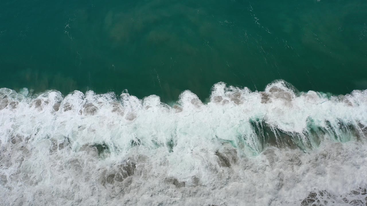 Overhead view of green ocean water in Los Cabos, Mexico, with a white surf band crossing the lower half and no visible people.