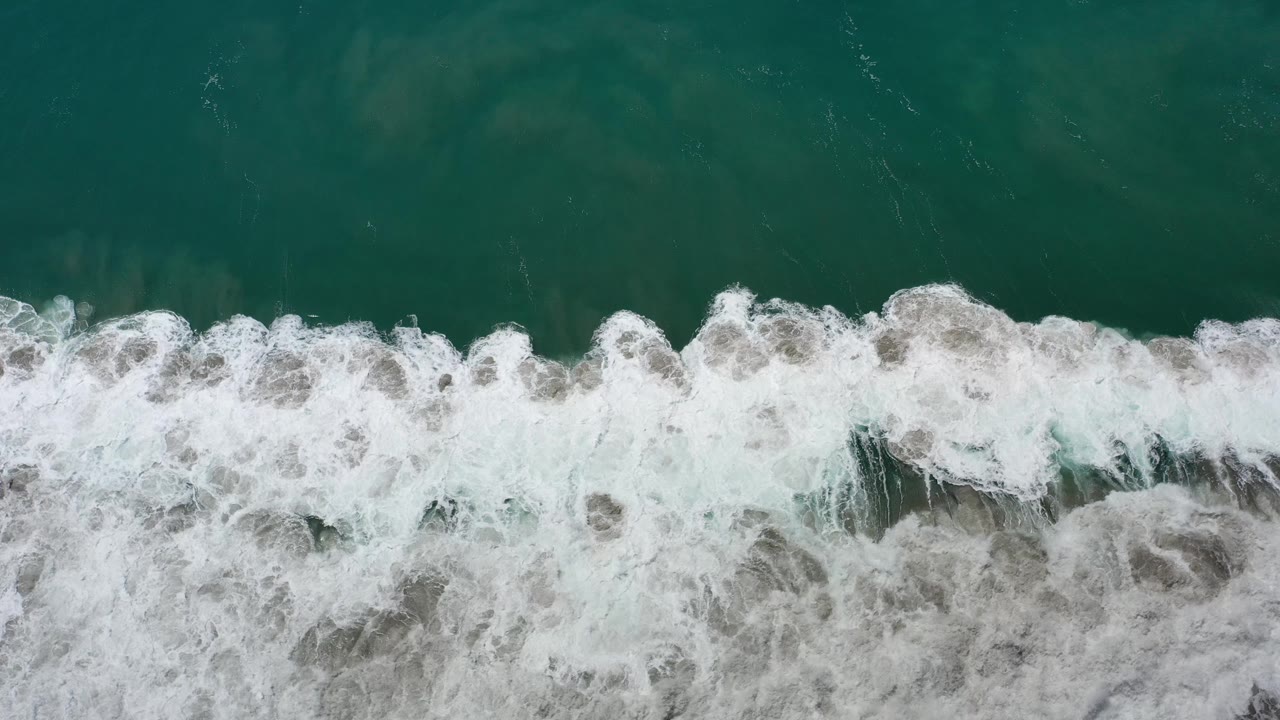 Aerial view of white surf breaking over darker green ocean water.