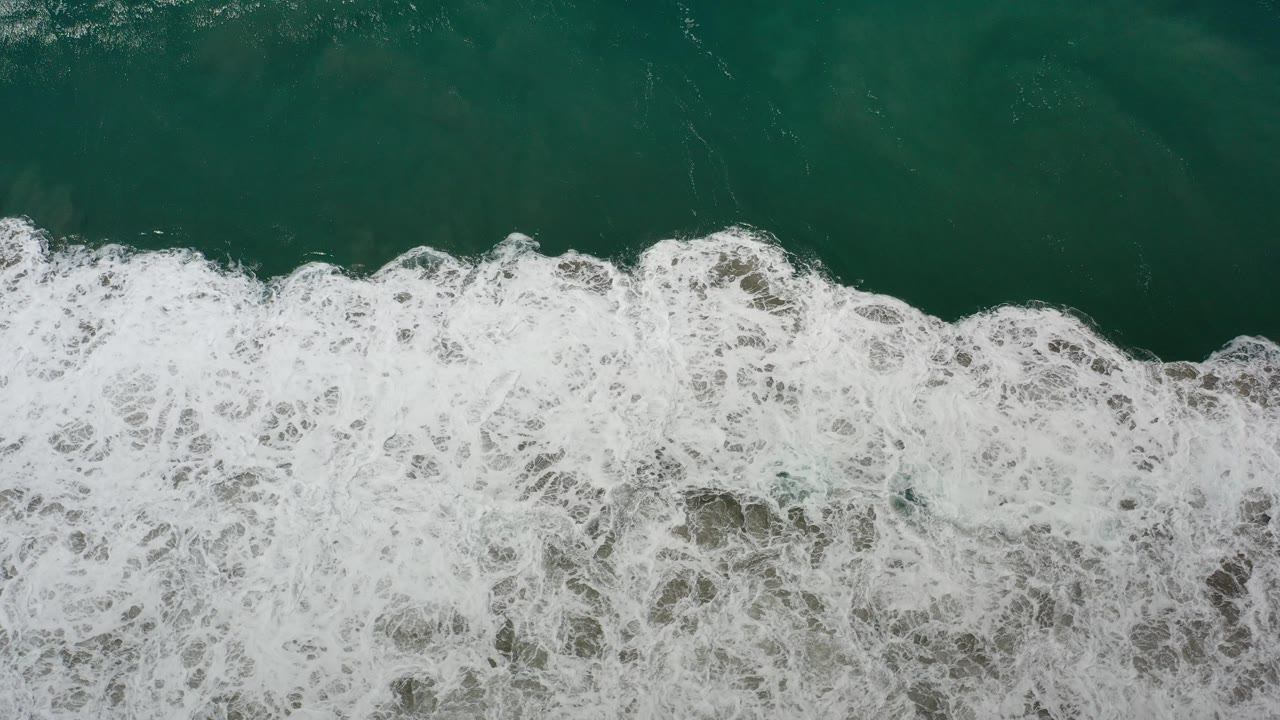 Overhead view in Los Cabos, Mexico, showing a horizontal band of white wave foam meeting dark green water, with no visible people.