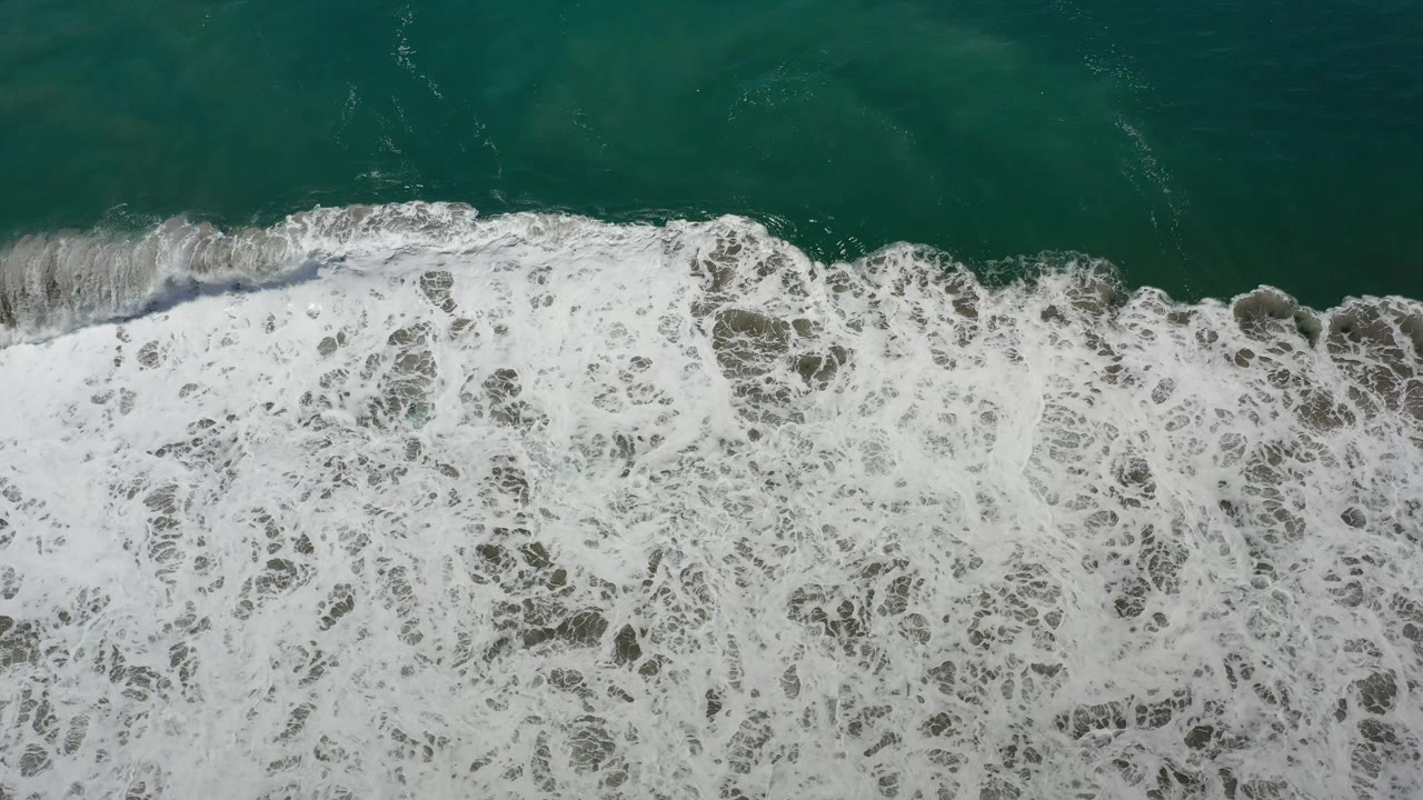 Overhead Los Cabos surf frame with white foam spreading across darker gray-green water below a teal ocean strip, with no visible people.