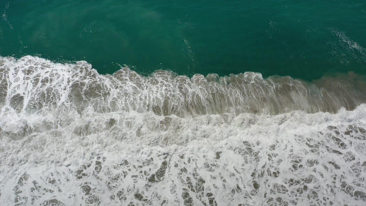 Overhead view of teal ocean water and white surf breaking onto a sandy beach in Los Cabos, Mexico, with no visible people.