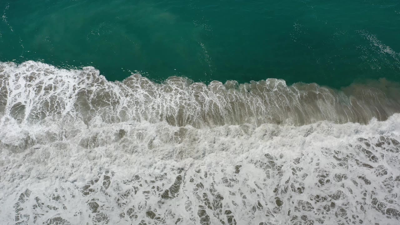 A band of white foam spreads across shallow green-blue water near the Los Cabos shoreline, with no visible people.