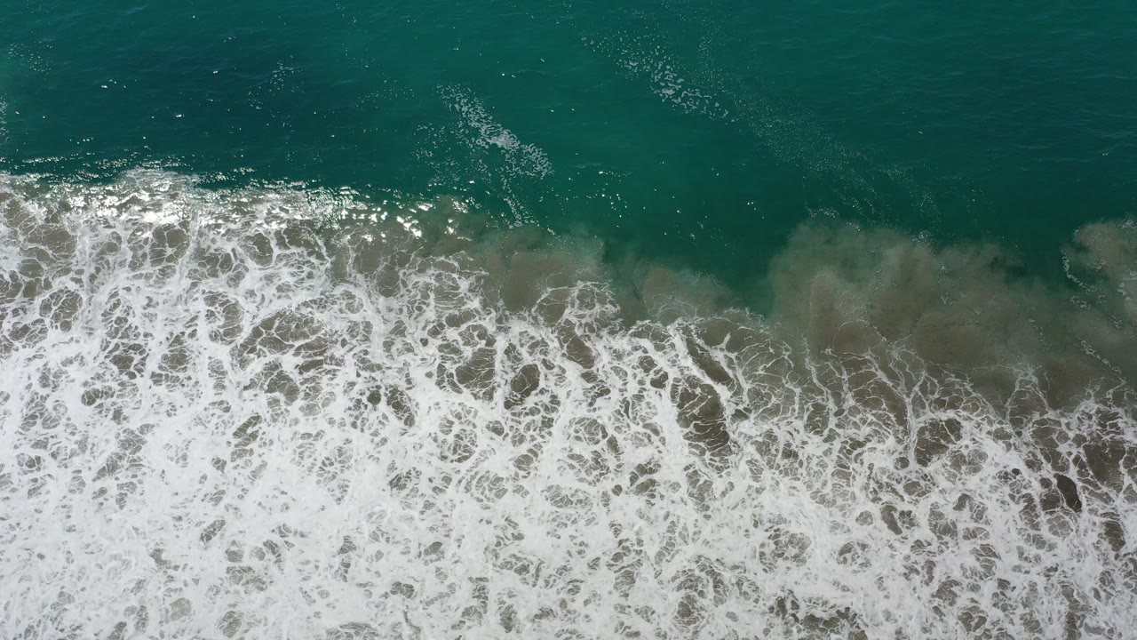Overhead view of green ocean water in Los Cabos, Mexico, with a broad band of white surf foam pushing up from the lower edge and no visible people.
