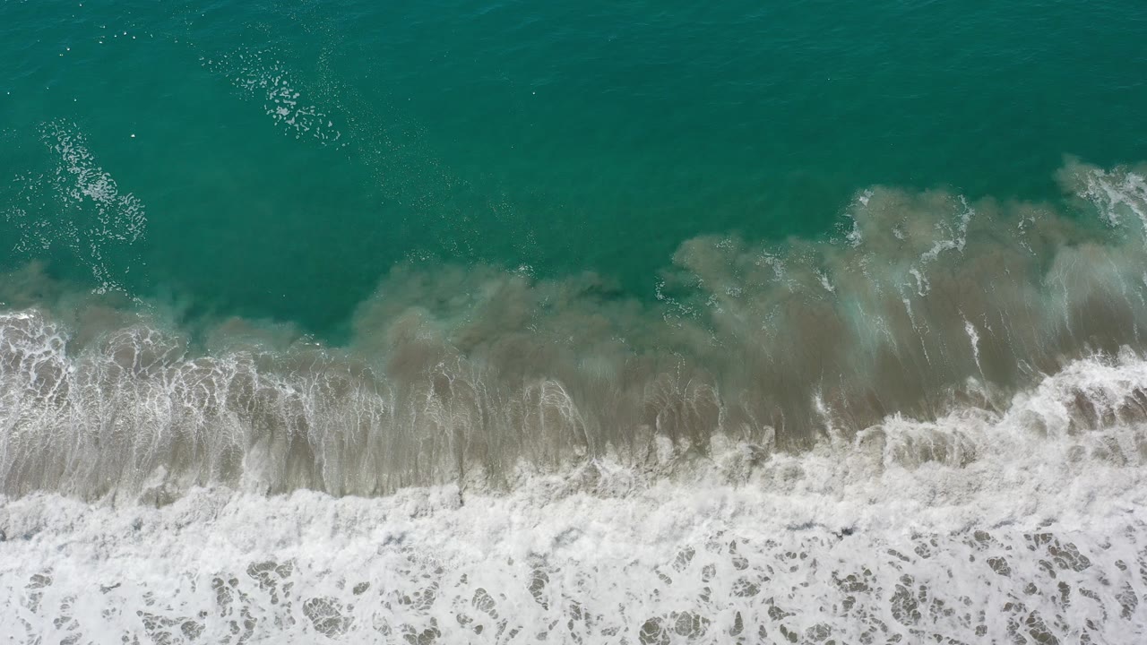 Overhead view in Los Cabos, Mexico of teal water meeting gray shallows and a band of white surf foam, with no visible people.