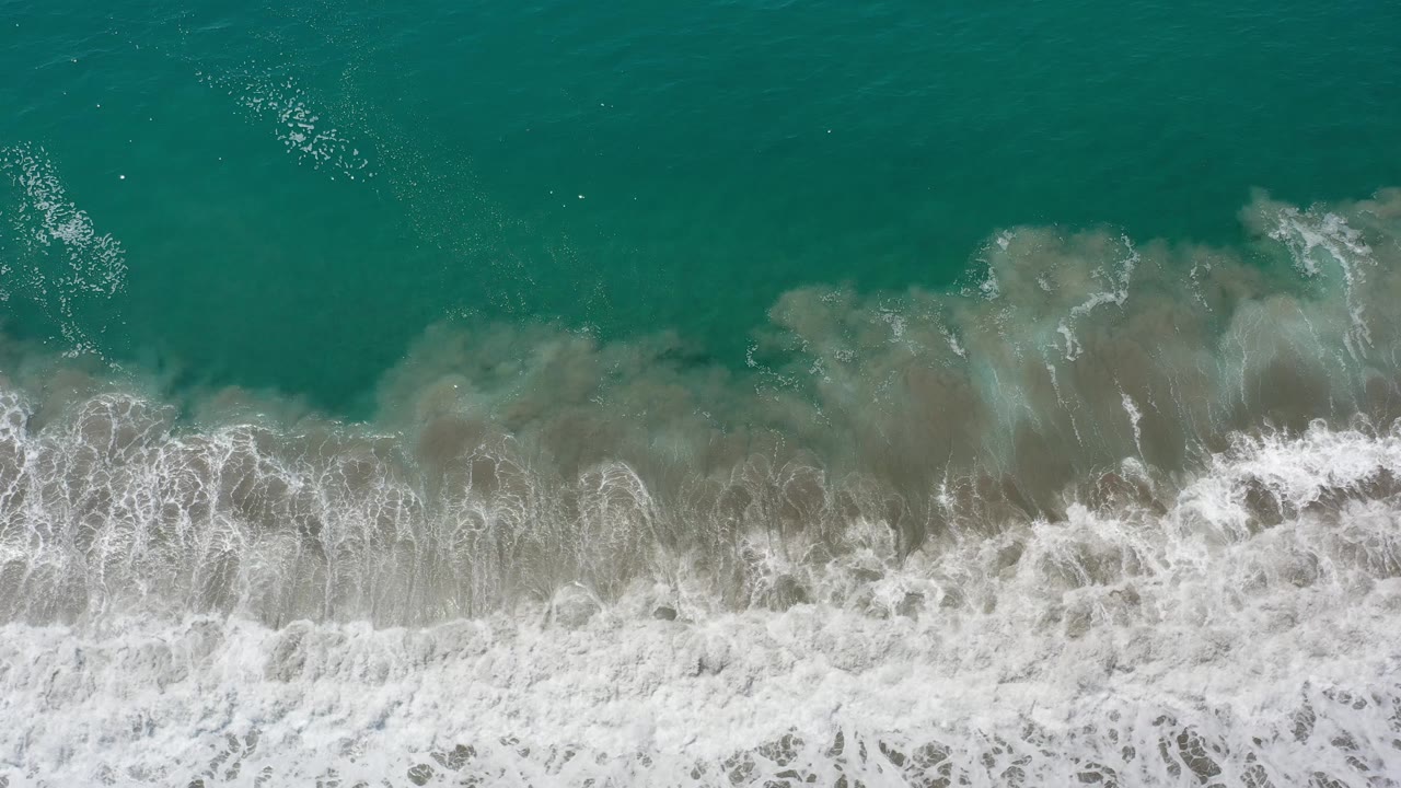 Aerial view of a breaking wave line with teal water above and foam below.