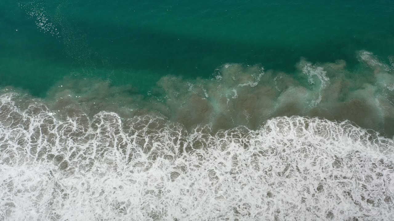 Overhead view of green ocean surf in Los Cabos, Mexico, with white foam clustered along the lower edge and no visible people.