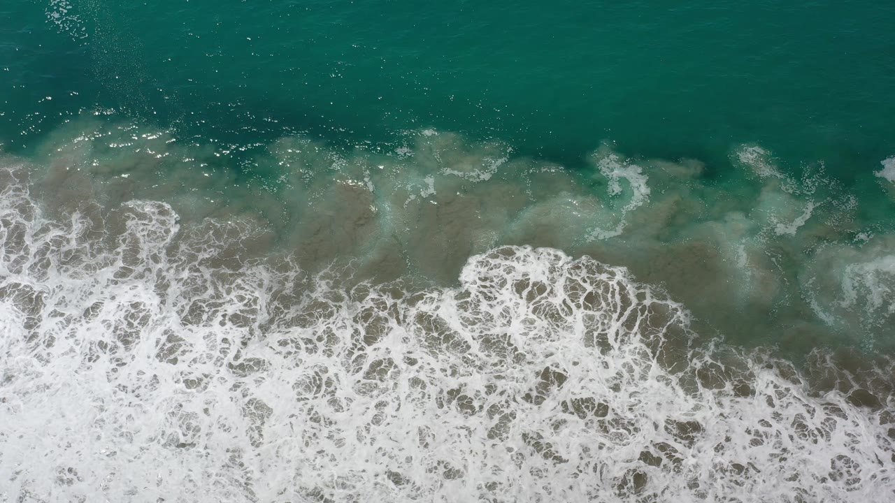 A dense band of white foam crosses the lower frame over green-blue water and submerged rock in Los Cabos, with no visible people.