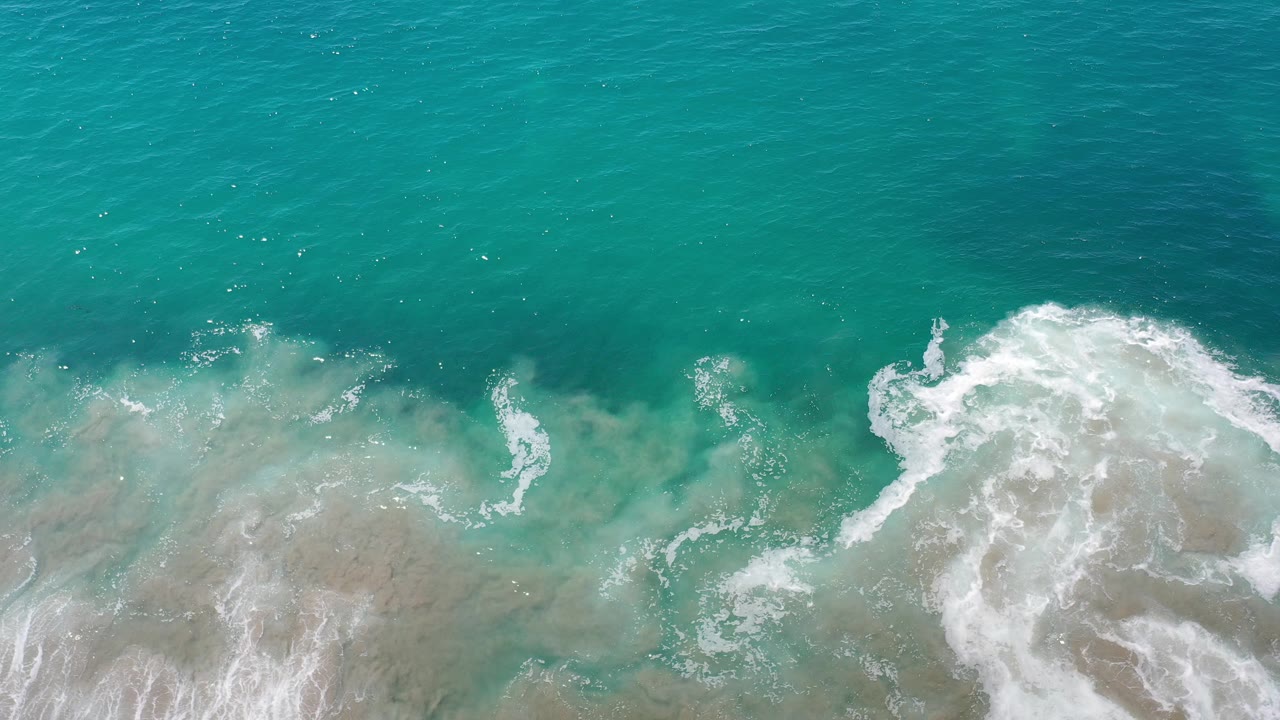 Overhead view of turquoise surf in Los Cabos, Mexico, with white foam spreading across pale shallow water and no visible people.
