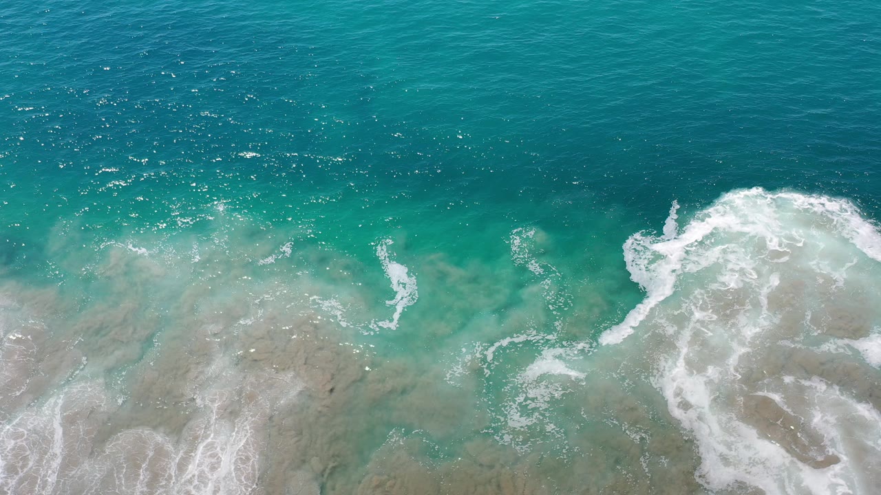 Overhead view of turquoise water and white surf washing over pale sand in Los Cabos, Mexico, with no visible people.