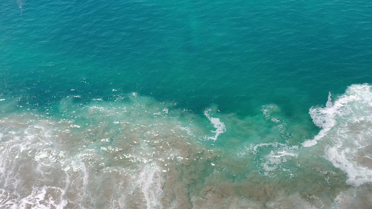 Overhead view of turquoise water and white surf foam rolling over shallow sand in Los Cabos, Mexico, with no visible people.