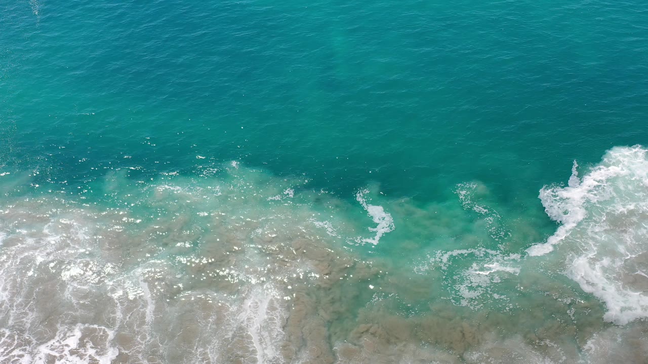 White wave foam spreads wider across translucent blue-green shoreline water in Los Cabos, Mexico, with no visible people.
