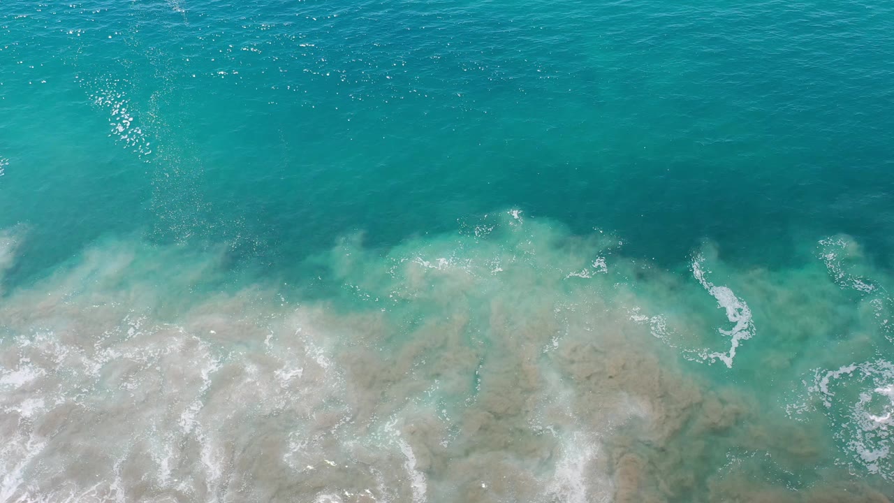 Aerial frame of blue-green ocean and foamy surf pushing across submerged sand in Los Cabos, Mexico, with no visible people.