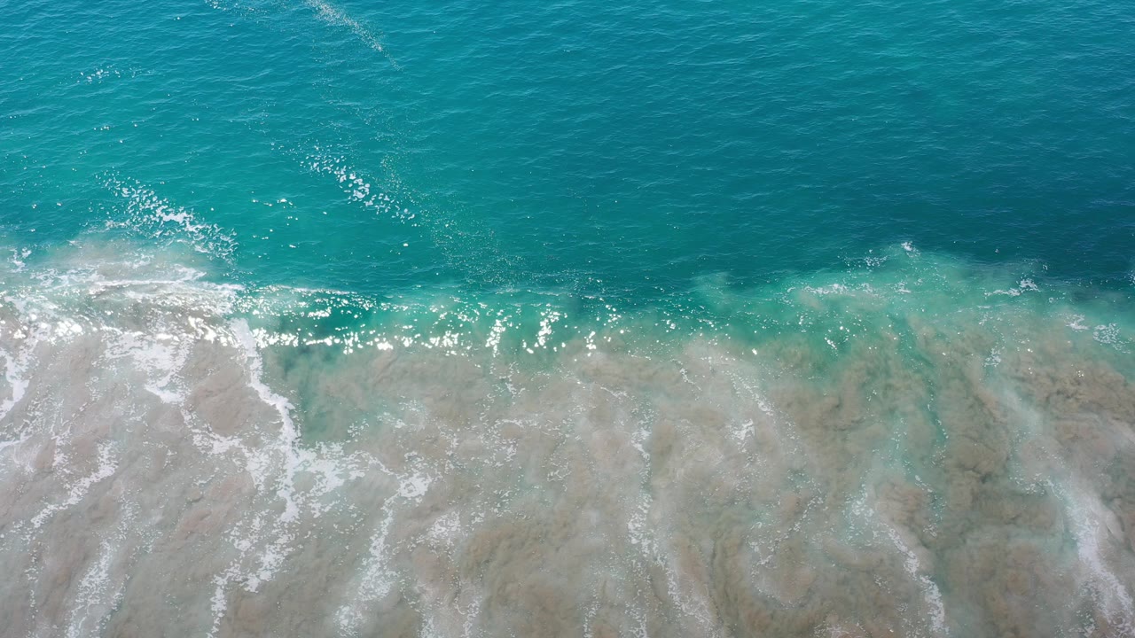 Overhead view of turquoise ocean water meeting wet sand in Los Cabos, Mexico, with scattered white foam and no visible people.