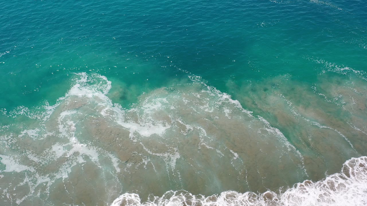 Opening overhead frame of blue-green ocean swell and white foam over shallow water in Los Cabos, Mexico, with no visible people.