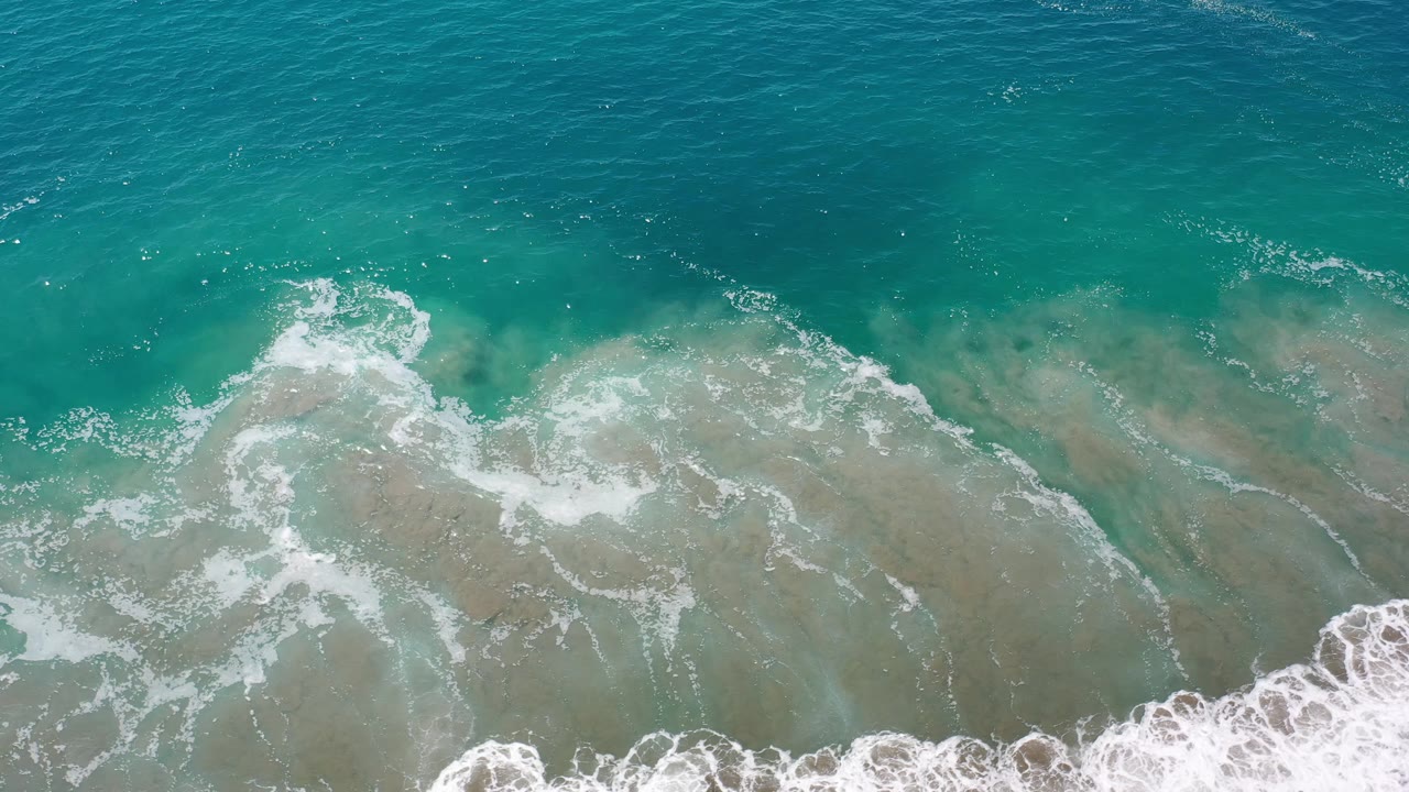 Overhead Los Cabos ocean frame with a larger white foam patch drifting across green-blue shallows and no visible people.