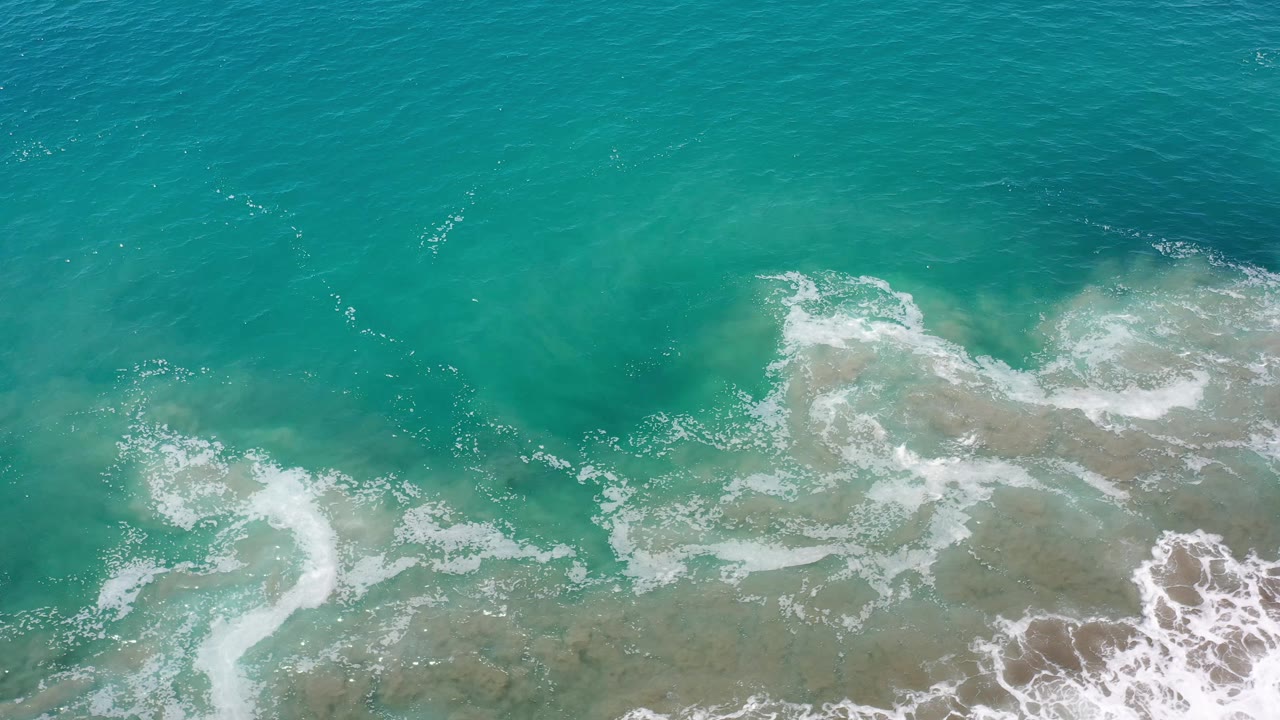 Top-down Los Cabos ocean frame with a curved white foam band at lower left, pale shallows through the center, and no visible people.