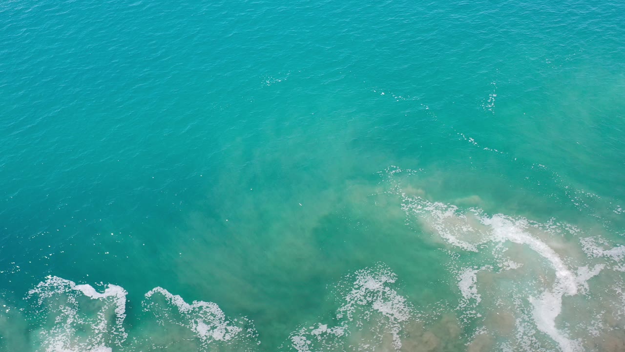 Top-down view of turquoise ocean in Los Cabos, Mexico, with white foam patches along the lower edge and no visible people.
