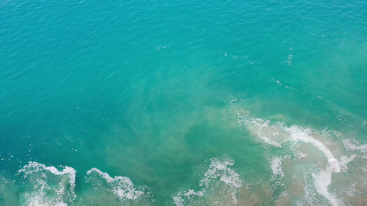 Aerial view over aqua water in Los Cabos, Mexico, showing pale sandy clouding beneath the surface and scattered foam, with no visible people.