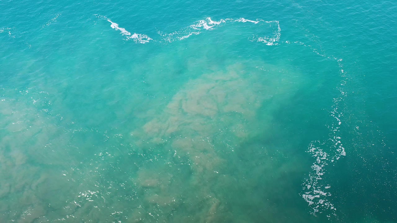 Overhead view of turquoise water in Los Cabos, Mexico, with pale underwater sand patches, white surf lines near the top, and no visible people.