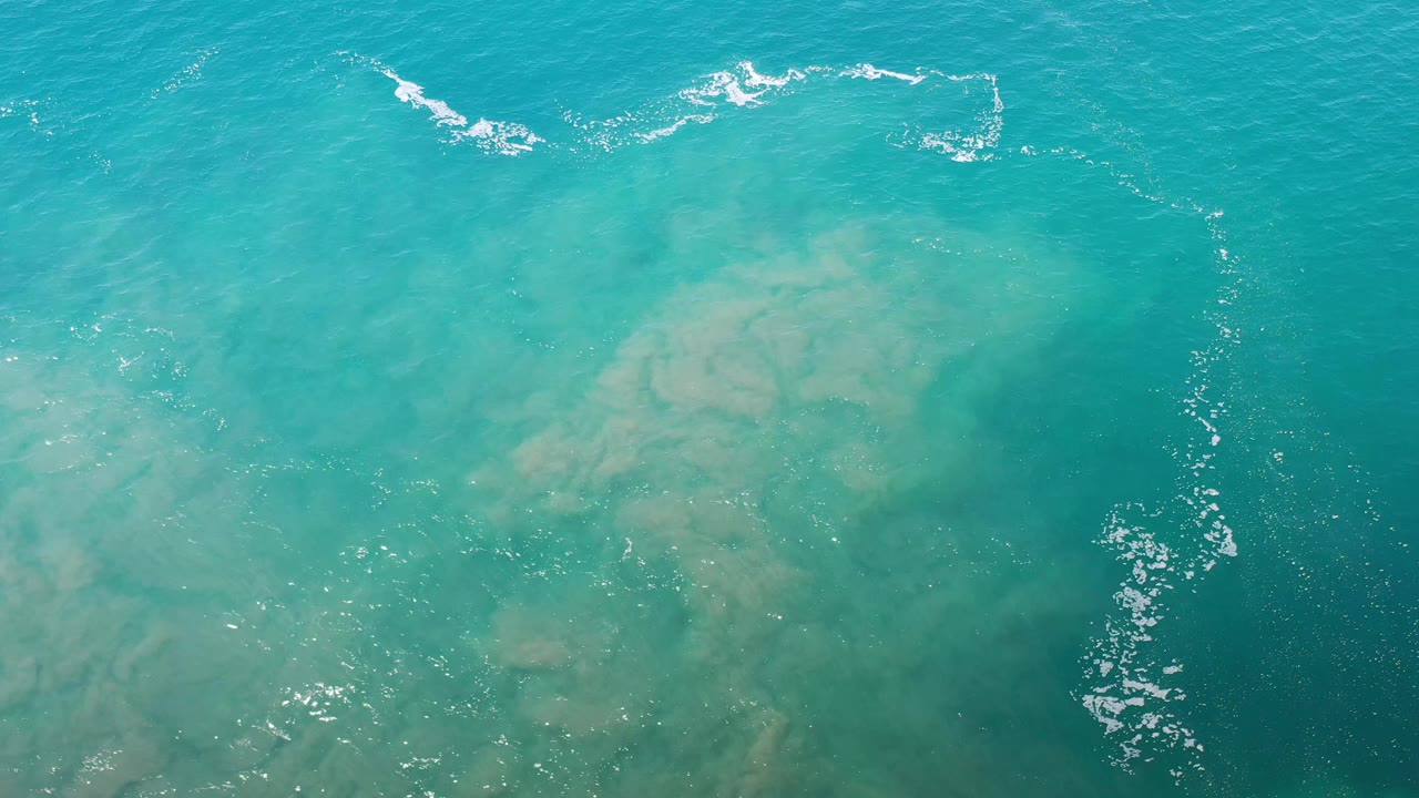 Aerial frame over Los Cabos water showing blue-green shallows, tan submerged sand forms, scattered sun sparkle, and no visible people.