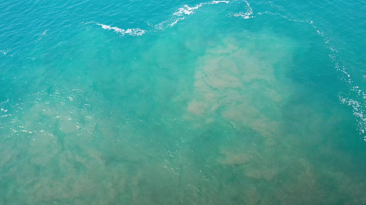 Overhead view of turquoise ocean water and tan underwater sand in Los Cabos, Mexico, with a short white wave line near the upper edge and no visible people.