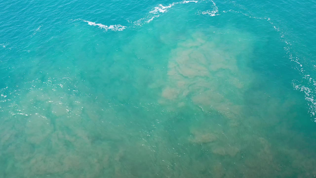 Blue-green water covers pale tan sand in Los Cabos, Mexico, with small white foam traces toward the upper right and no visible people.