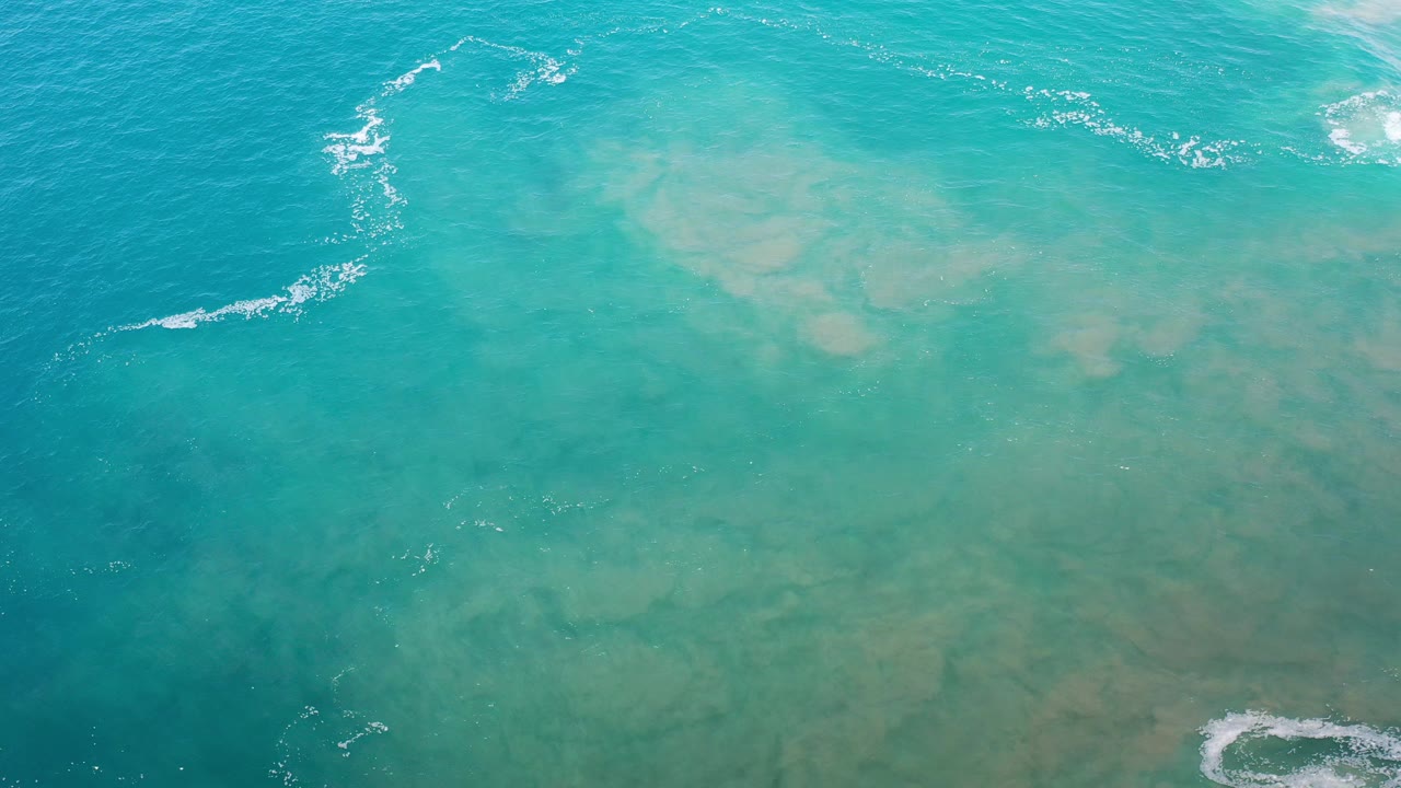 Overhead view of turquoise and green sea in Los Cabos, Mexico, with pale underwater patches, small white wave streaks near the top, and no visible people.