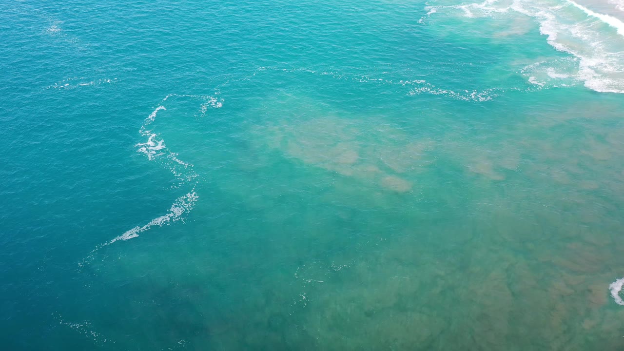Aerial frame over Los Cabos water showing teal ocean, muted sandbars below the surface, scattered white foam, and no visible people.