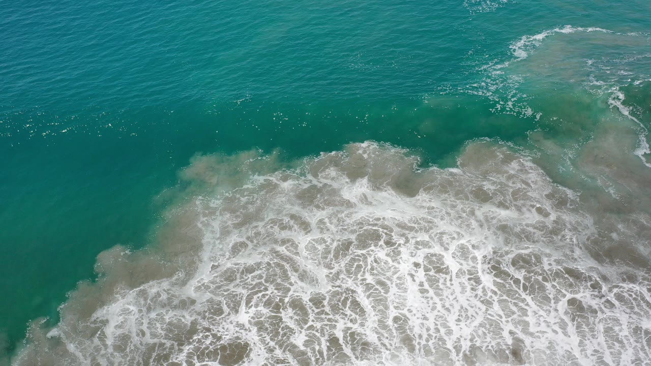 Overhead view of green ocean surf in Los Cabos, Mexico, with white foam spreading across shallow water and no visible people.