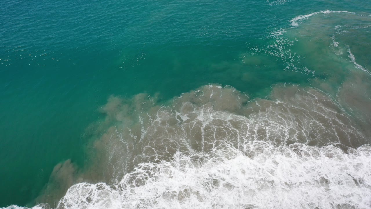 Overhead view of teal water and white surf washing across shallow sand in Los Cabos, Mexico, with no visible people.