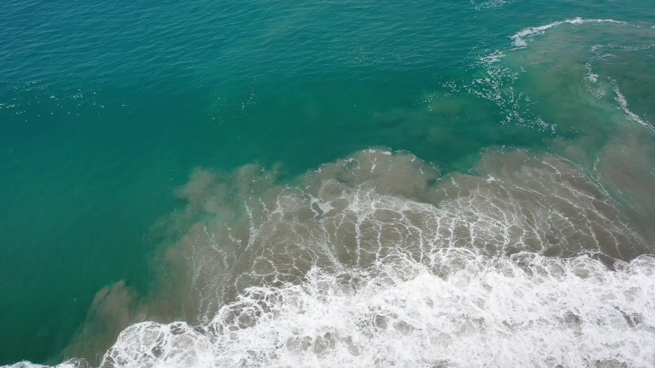 Aerial frame of foamy tide patterns spreading over pale underwater sand in Los Cabos, Mexico, with no visible people.