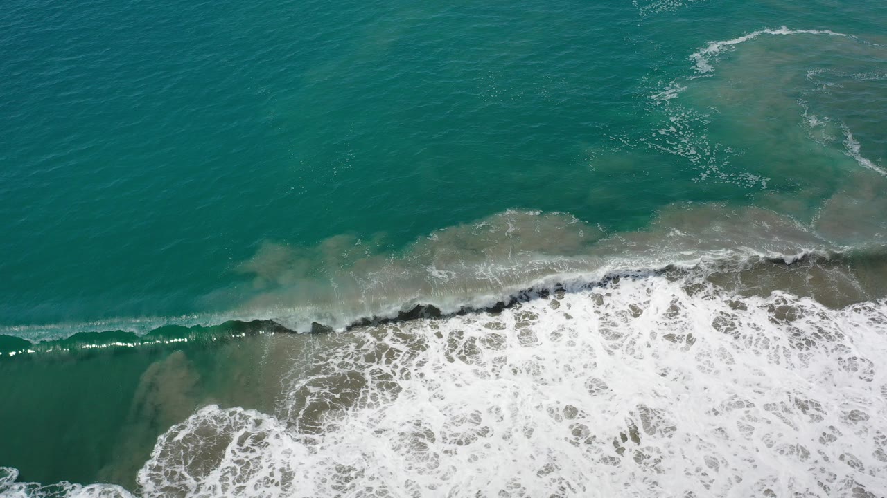 Overhead view in Los Cabos, Mexico, with turquoise ocean above a band of white surf breaking over tan sand and no visible people.