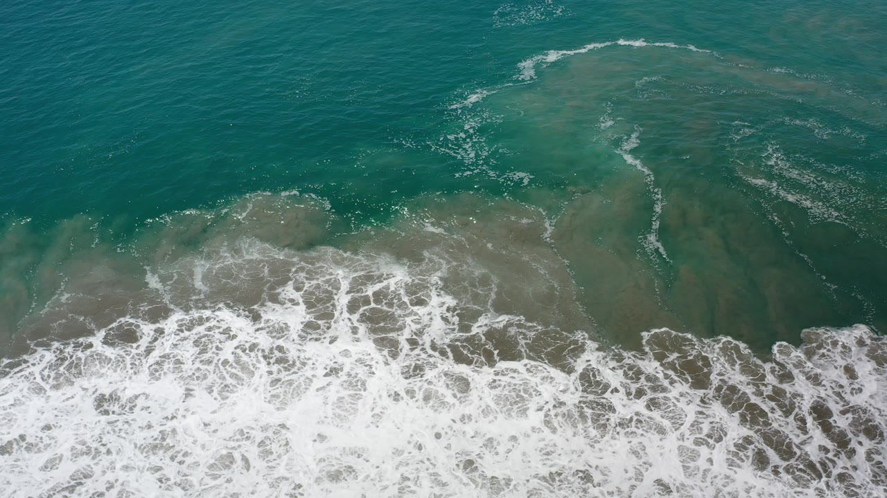 Top-down view of green-blue ocean water and white shore break in Los Cabos, Mexico, with no visible people.