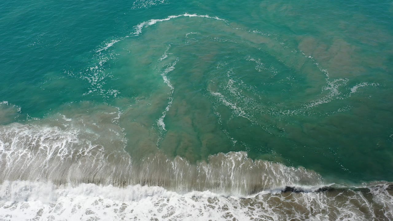 Overhead view of aqua surf and sandy shallows in Los Cabos, Mexico, with white foam crossing the lower edge and no visible people.