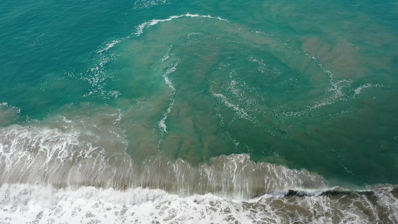 A high overhead Los Cabos beach view shows translucent turquoise water over sand, with a curving foam line offshore and no visible people.
