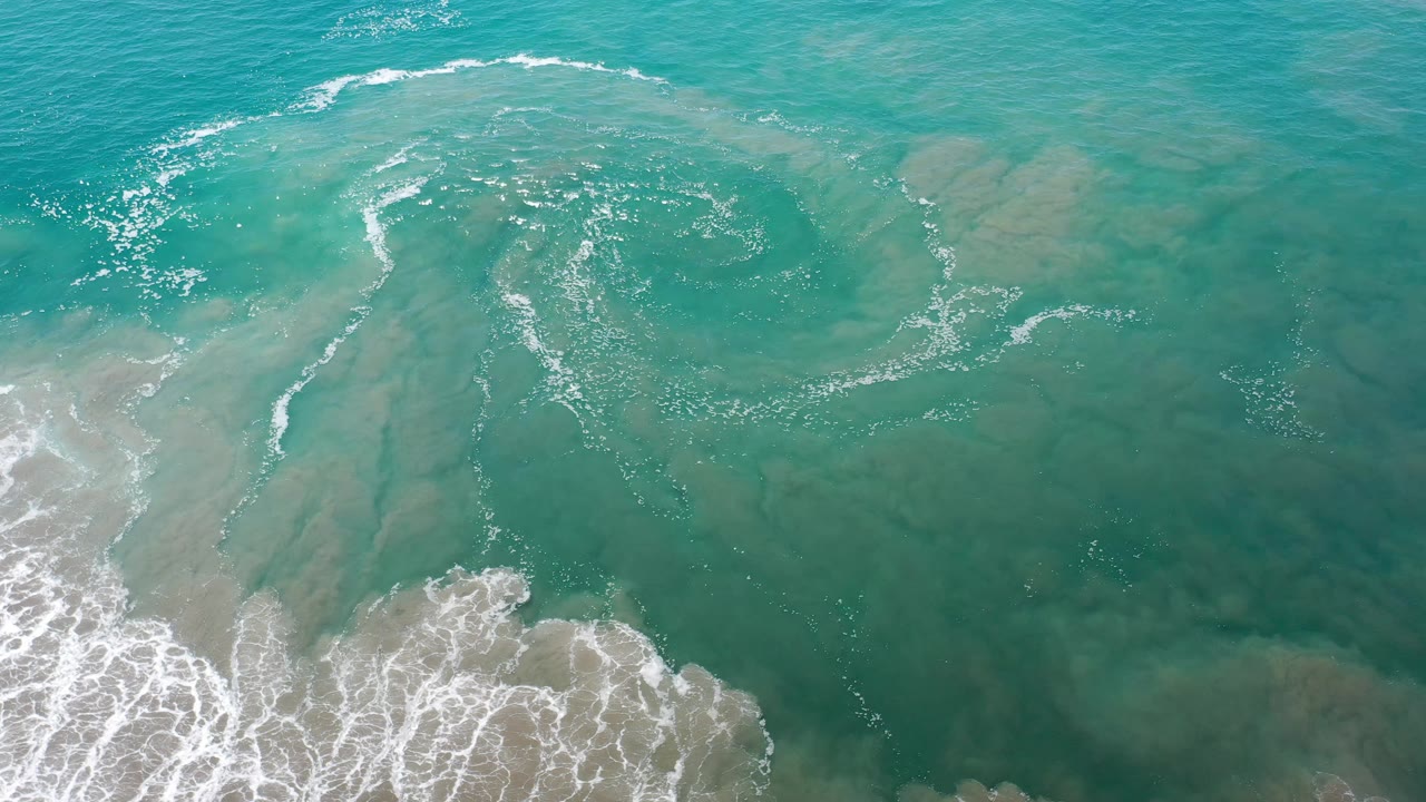 Aerial frame of Los Cabos teal water with pale sandy areas visible under the surface and a broken curve of white foam across the upper half, no visible people.