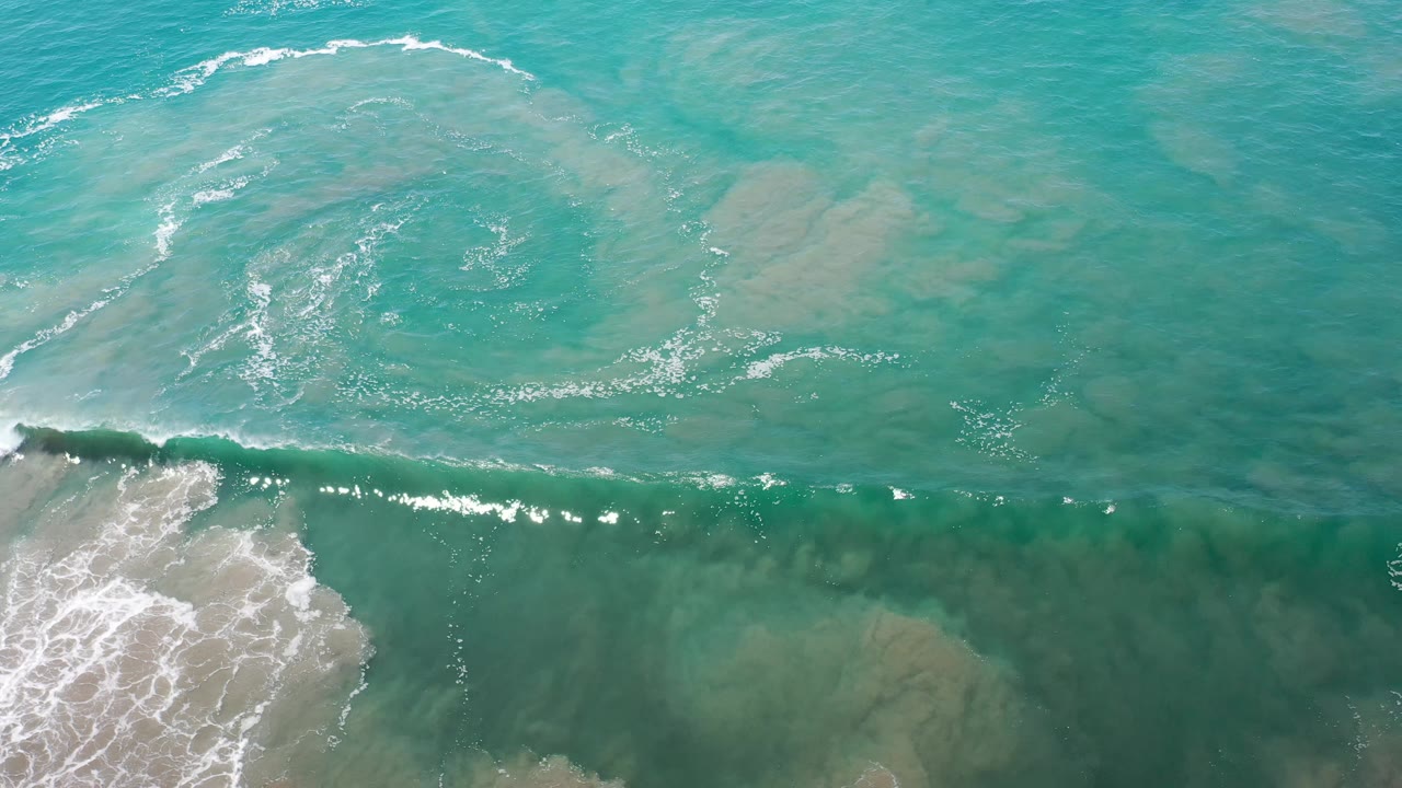 Overhead Los Cabos ocean frame with clear turquoise water, scattered pale underwater patches, a curved foam pattern, and no visible people.
