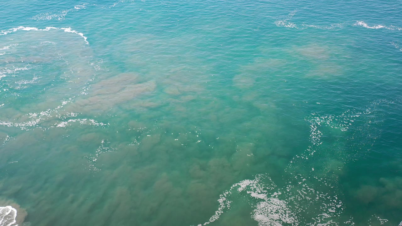 Aerial view over Los Cabos, Mexico water, with turquoise shallows, darker green patches, white foam near the top and lower left, and no visible people.