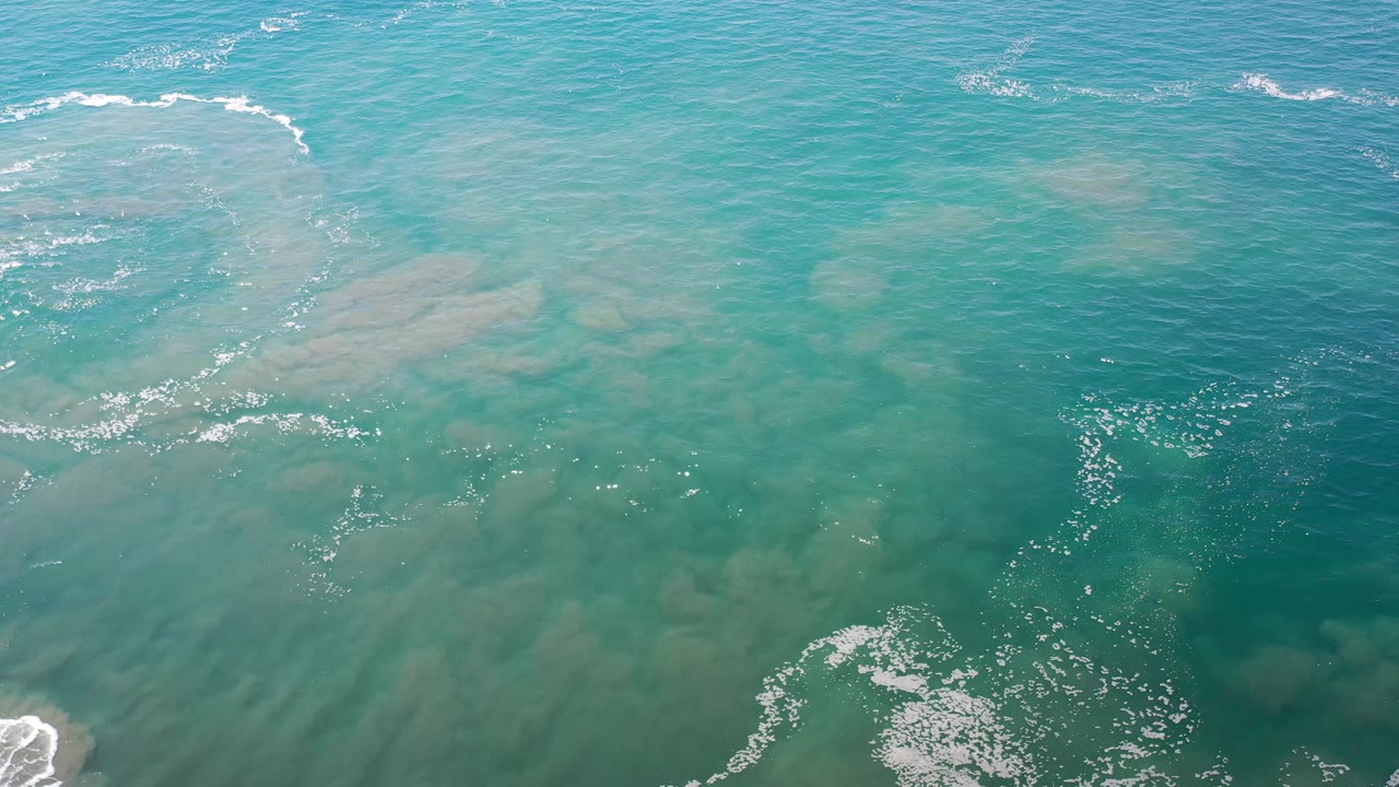 Overhead Los Cabos, Mexico ocean view with a curved white foam break near the upper center, sandy green water below, and no visible people.
