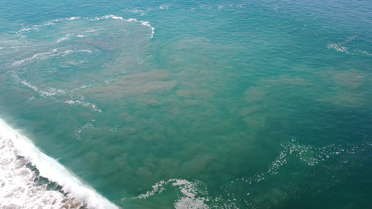 Overhead Los Cabos sea view with pale underwater sand shapes, scattered foam, and a breaking wave along the left edge with no visible people.