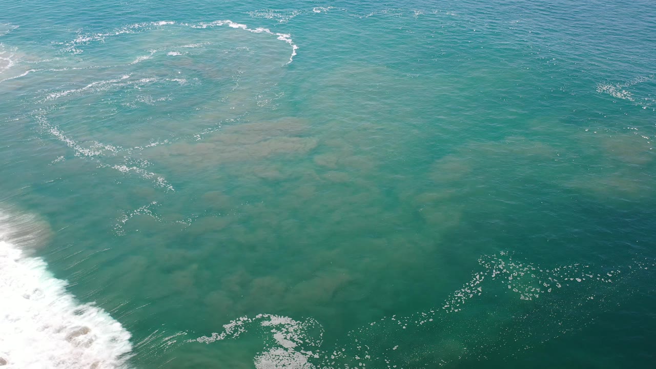 Overhead ocean view with white wave wash at left and darker green water at bottom right.