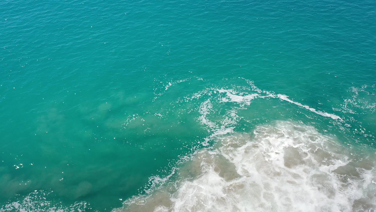 Overhead view of turquoise shallow water in Los Cabos, Mexico, with white foam entering from the lower frame and no visible people.