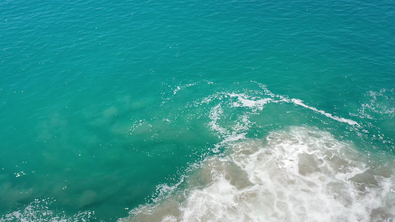 Overhead Los Cabos water view with a larger patch of white foam spreading across submerged sand and no visible people.