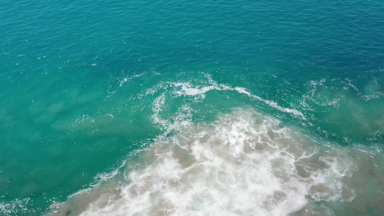 Overhead view in Los Cabos, Mexico where foamy surf curls across blue-green water, with the brightest whitewater concentrated near the lower right and no visible people.