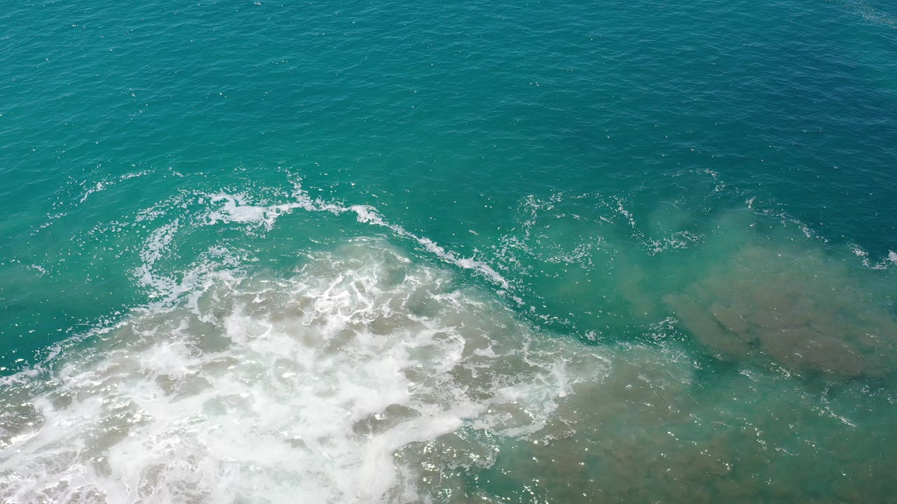 Aerial view of turquoise ocean surf over shallow rocky water.