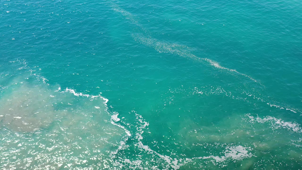 Blue-green ocean surface in Los Cabos, Mexico, with a foamy wave edge crossing sandy water and no visible people.